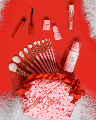 Rózsaszín karácsonyi P.Louise sminktáska piros, ünnepi dizájnnal, sminktermékekkel, köztük sminkecsettel, ajakolajjal, szempillaspirállal és egyebekkel, piros háttéren hóval.