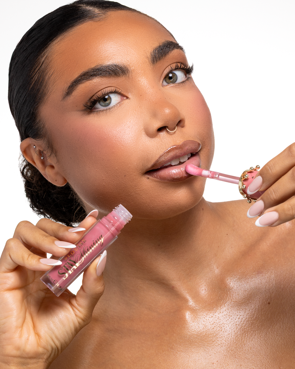 Közeli kép egy nőről, aki egy rózsaszín P.Louise Stay Shining Lip Oil folyékony ajakterméket alkalmaz egy őzike-talp applikátorral, miközben a másik kezében tartja a csövet, tiszta fehér háttér előtt.