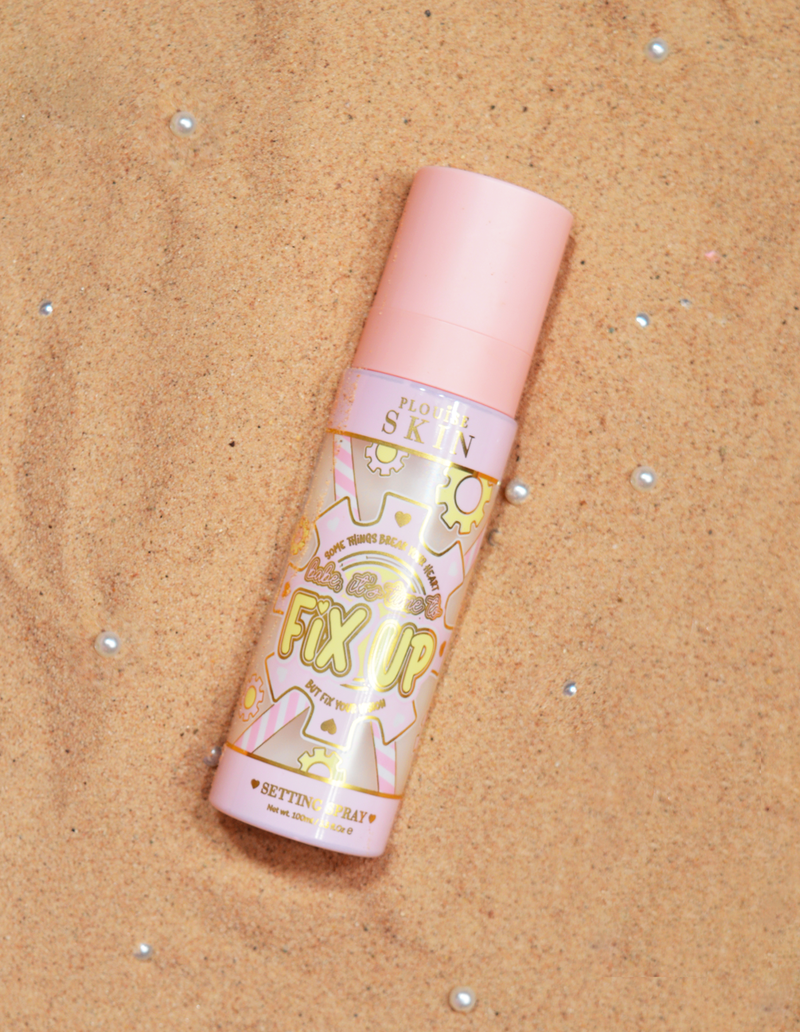 Egy üveg P.Louise Fix Up Mattifying Setting Spray rózsaszín kupakkal és dekoratív mintákkal a címkén, homokos felületen.