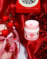 A P.Louise Plot Twist Make It Melt sminktisztító balzsam tartálya vörös szövetfelületen díszített elemekkel, köztük egy cseresznyével és egy vintage piros telefonnal a háttérben.