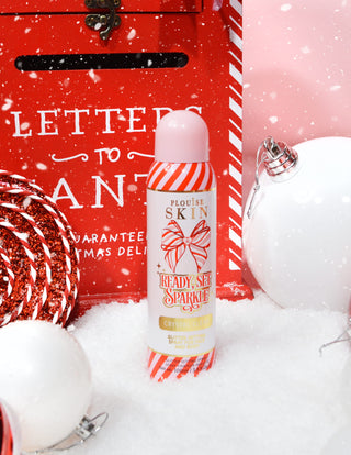 Egy üveg P. Louise Ready Set Sparkle Christmas Glitter Setting Spray olyan ünnepi díszekkel körülvéve, mint a hó, csecsebecse és egy karácsonyi levélszekrény